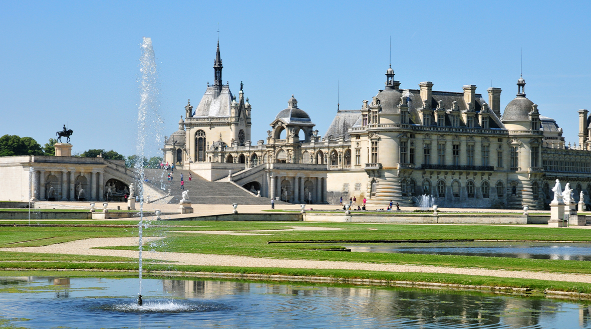 A fountain sprays water in the gardens of a lavish country house, with a château in the background